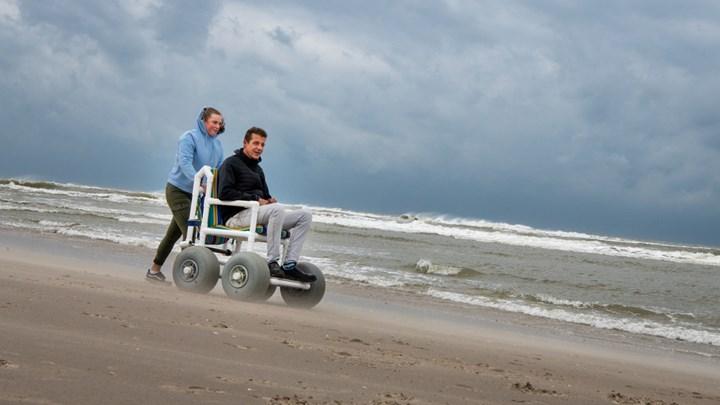 Vrouw duwt man voort op het strand in een jutter, een strandrolstoel.